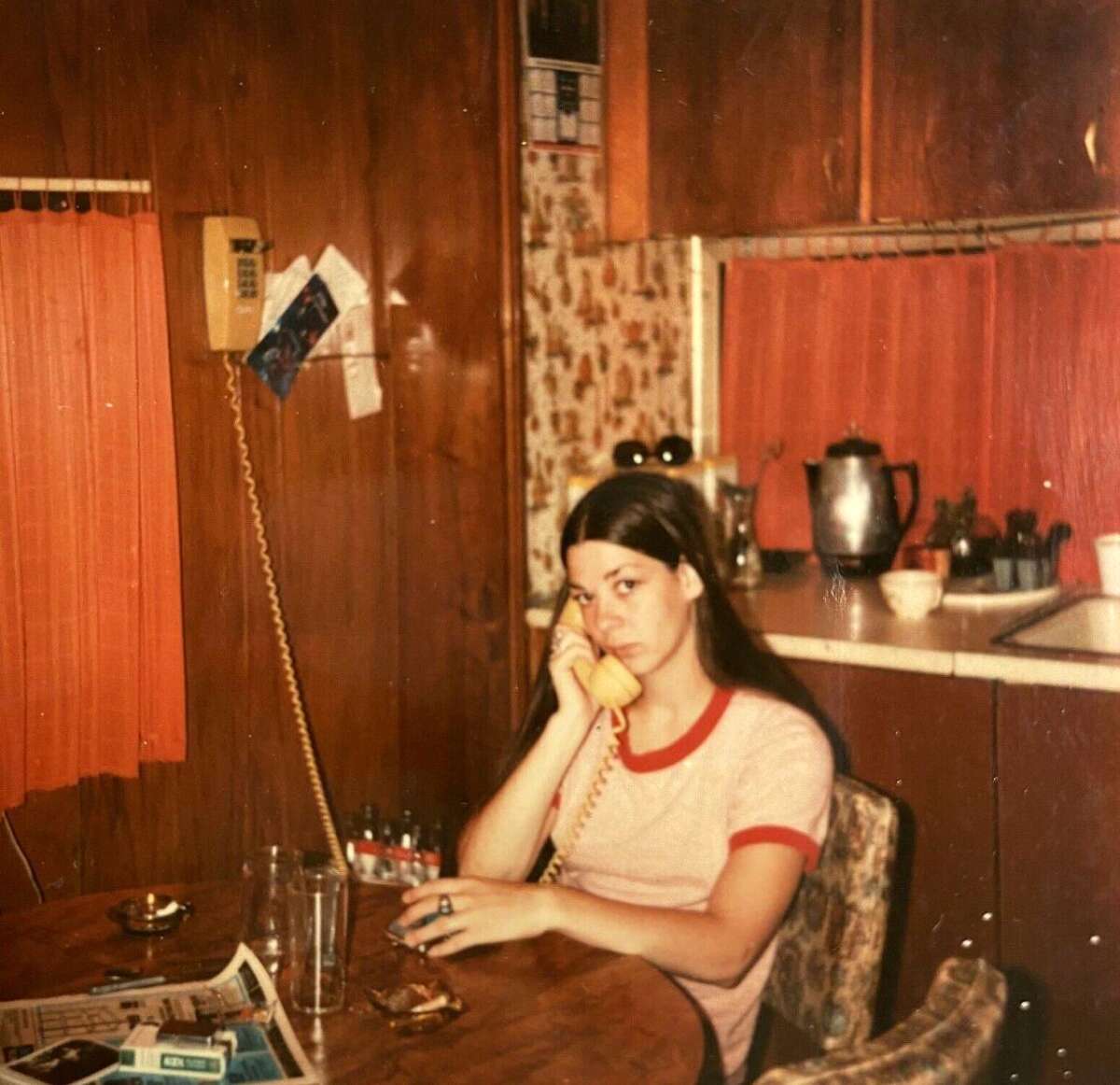 A young woman with long dark hair sits at a wooden table in a retro kitchen, talking on a yellow rotary phone. She wears a white and red ringer T-shirt and looks slightly serious. The kitchen has wood-paneled walls and orange curtains.