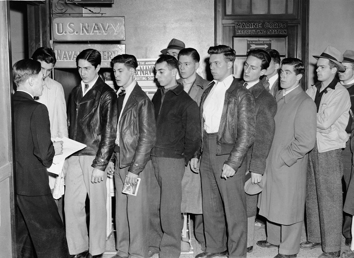 A group of young men wait in line at a U.S. Navy and Marine Corps recruiting station, many wearing jackets and holding papers, while an official stands at the front speaking to them.
