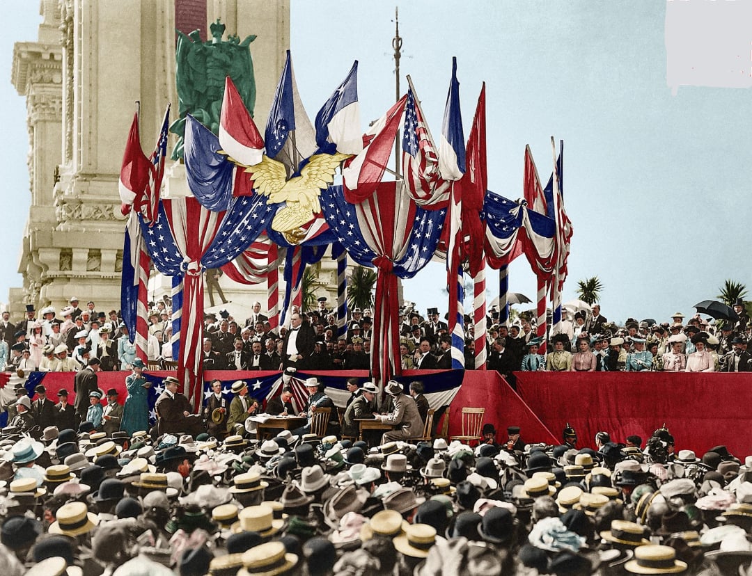 A large crowd gathers at an outdoor event, facing a decorated stage with red, white, and blue bunting, flags, and an eagle emblem. People in early 20th-century attire watch speakers at the podium.