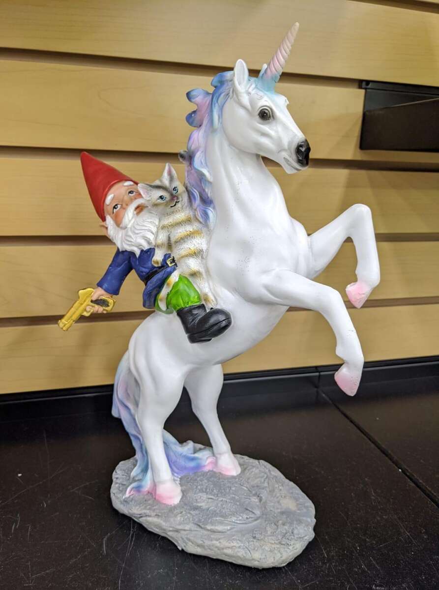 A figurine of a bearded gnome holding a toy gun and a grey tabby cat, riding a rearing white unicorn with a pink and blue mane, set against a wood-paneled background.