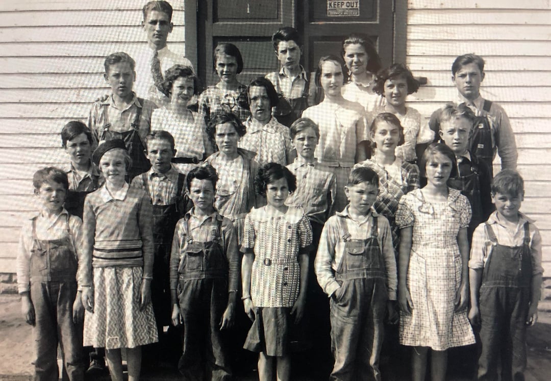 A black-and-white photo of a group of school children and one adult standing in rows outside a wooden building. Most children wear plaid shirts and overalls or dresses. A sign on the door behind them reads "KEEP OUT.