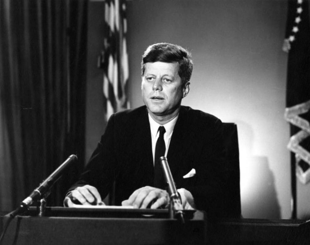 A man in a suit sits at a desk with microphones in front of him, appearing to speak or address an audience. American flags are visible in the background.