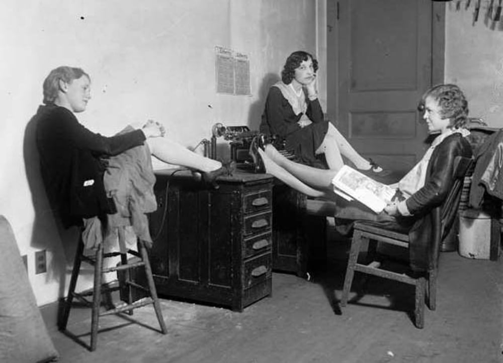 Three women sit and relax in a small room with their feet up on a desk. One reads a magazine, another leans back on a stool, and the third sits in the background with her hand on her chin. A typewriter is on the desk.
