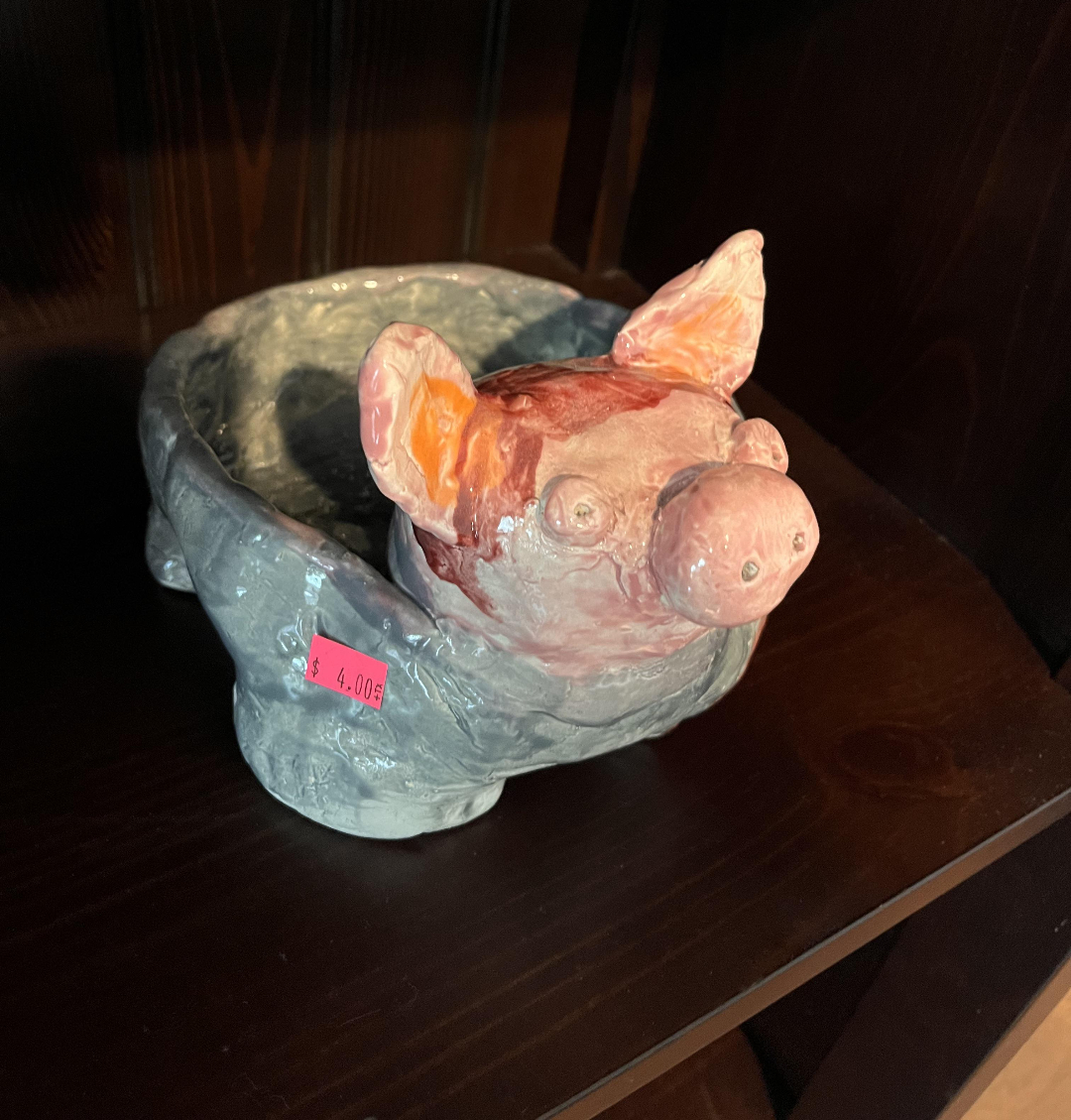 A handmade ceramic bowl shaped like a pig with large ears sits on a dark wooden shelf. The bowl has a pink price sticker showing $4.00 attached to its side.