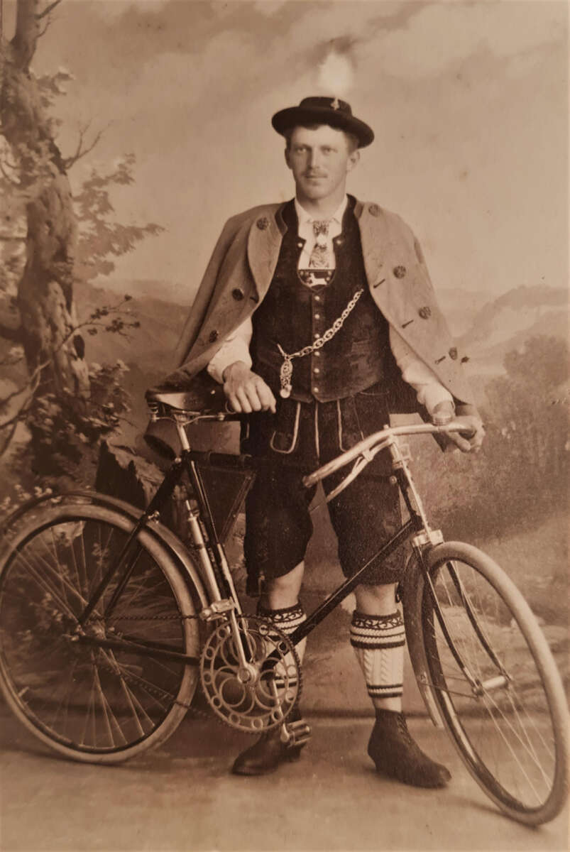 A man in traditional Bavarian attire stands beside a vintage bicycle, posing for a sepia-toned portrait in front of a painted nature backdrop.