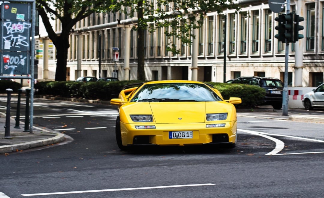 A bright yellow Lamborghini sports car drives around a curve on a city street, surrounded by buildings and trees, with a traffic light and some parked cars visible in the background.