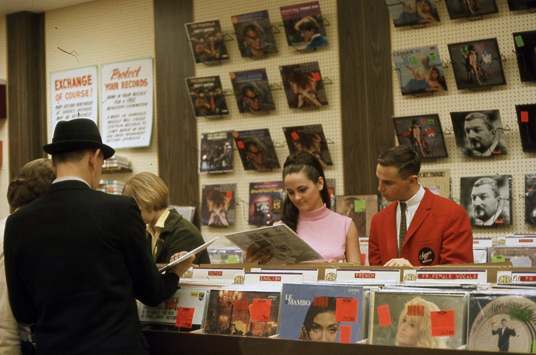 People browse and examine records in a vintage record store, with albums displayed on shelves and counters. A man in a red jacket assists a woman in pink, while others look through the vinyl selection.