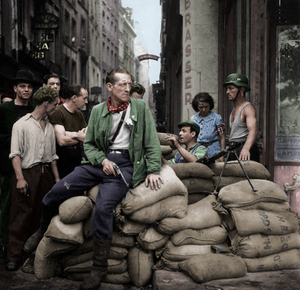 A group of people, some armed and in uniform, gather around sandbags on a city street, suggesting a wartime barricade. One man sits on the sandbags, while others stand or sit nearby, conversing.