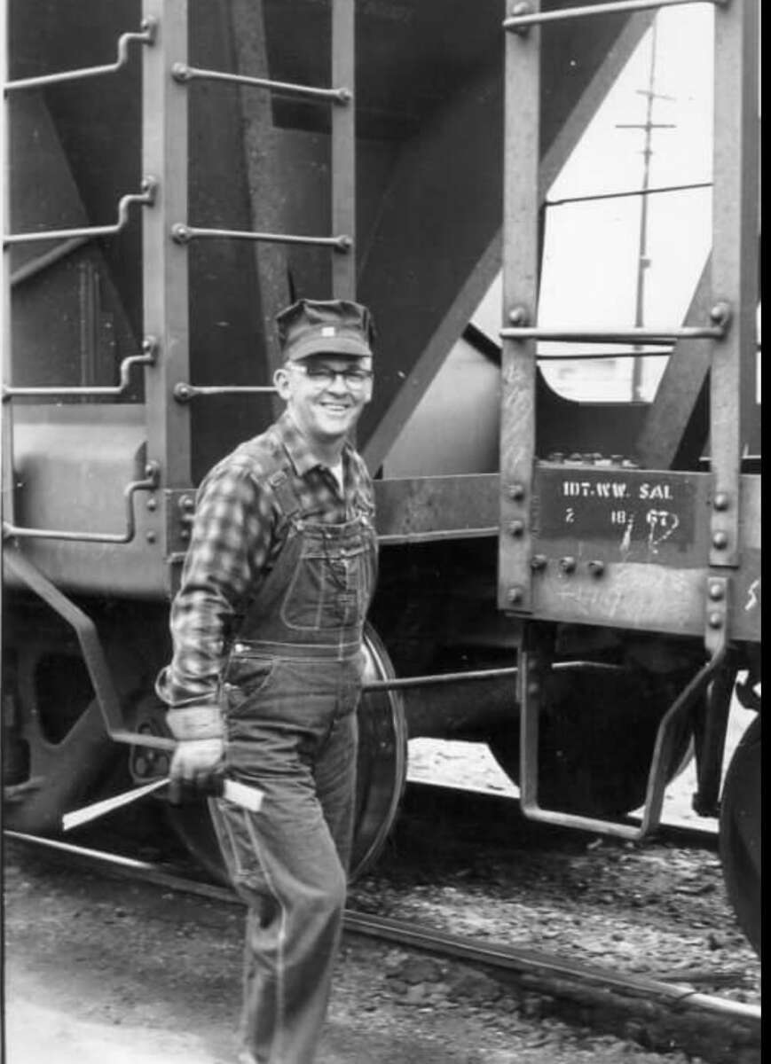 A man wearing a cap, glasses, plaid shirt, and overalls stands smiling in front of a train car on railroad tracks, holding a tool in his hand.