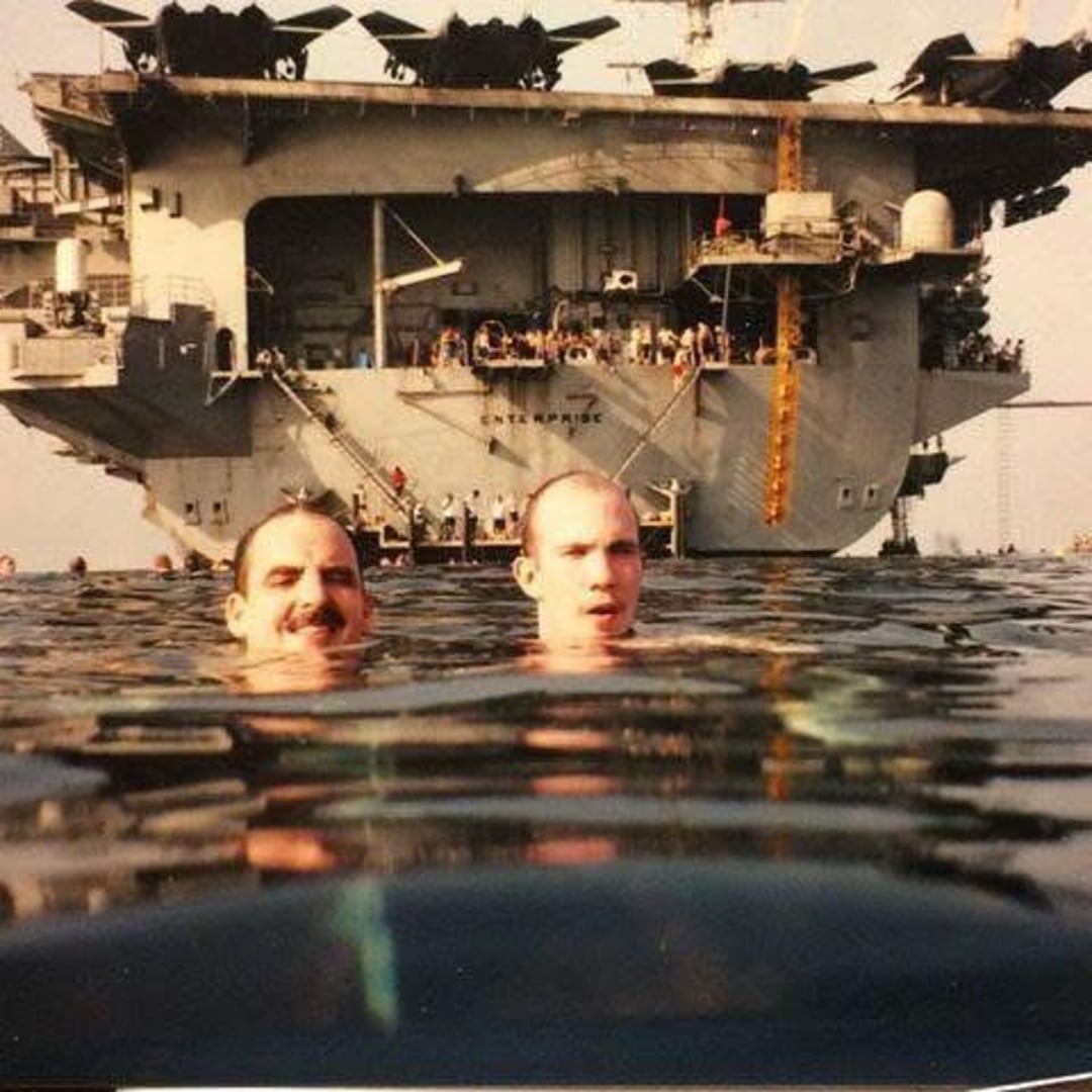 Two men swim in the ocean with their heads above water. Behind them, a large aircraft carrier labeled "Enterprise" is visible, with people standing on its deck and aircraft parked on top.