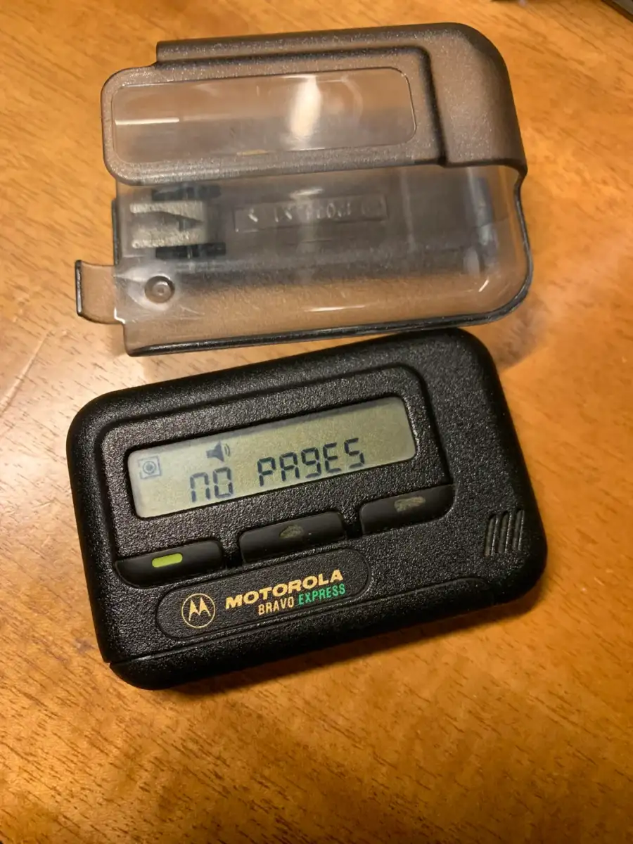A black Motorola Bravo Express pager with its clear plastic cover removed rests on a wooden surface. The pager’s screen displays the message “NO PAGES.”