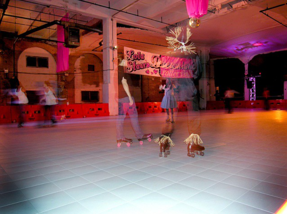 People roller skating in a large indoor rink with colorful lights, blurred motion suggesting movement. Pink banners hang from the ceiling, and a disco ball is visible above the rink.