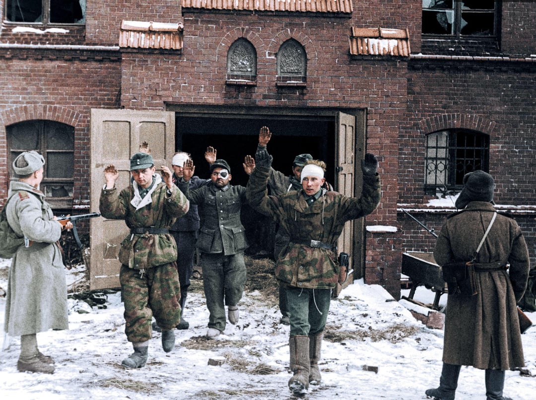 A group of soldiers with raised hands, surrendering to armed guards outside a brick building in snowy conditions. The guards point rifles at the prisoners, and broken windows are visible above the doorway.