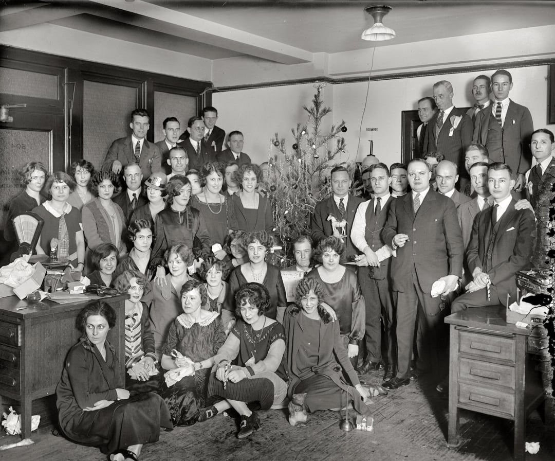 Black-and-white photo of a large group of men and women in formal attire gathered around decorated Christmas trees in an office, posing and smiling for the camera. Desks and paperwork are visible in the background.