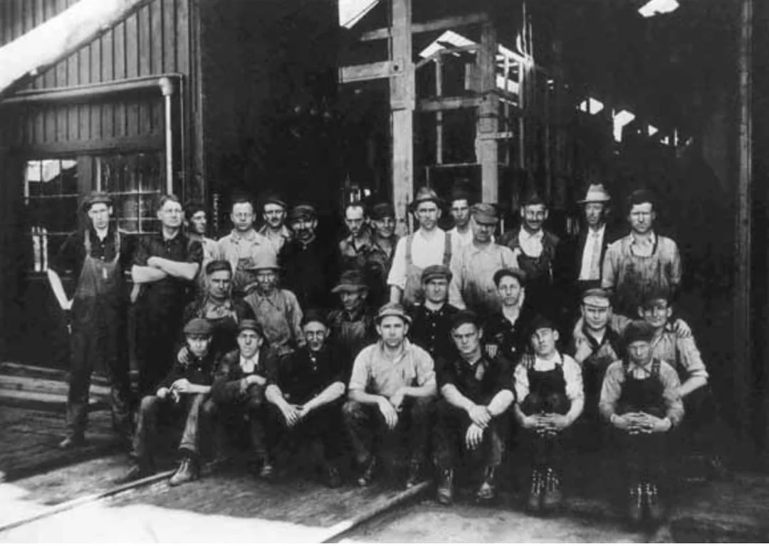 A black-and-white photo of about 25 men, some standing and some crouched, posing in front of a large industrial building or factory, wearing work clothes typical of the early 20th century.