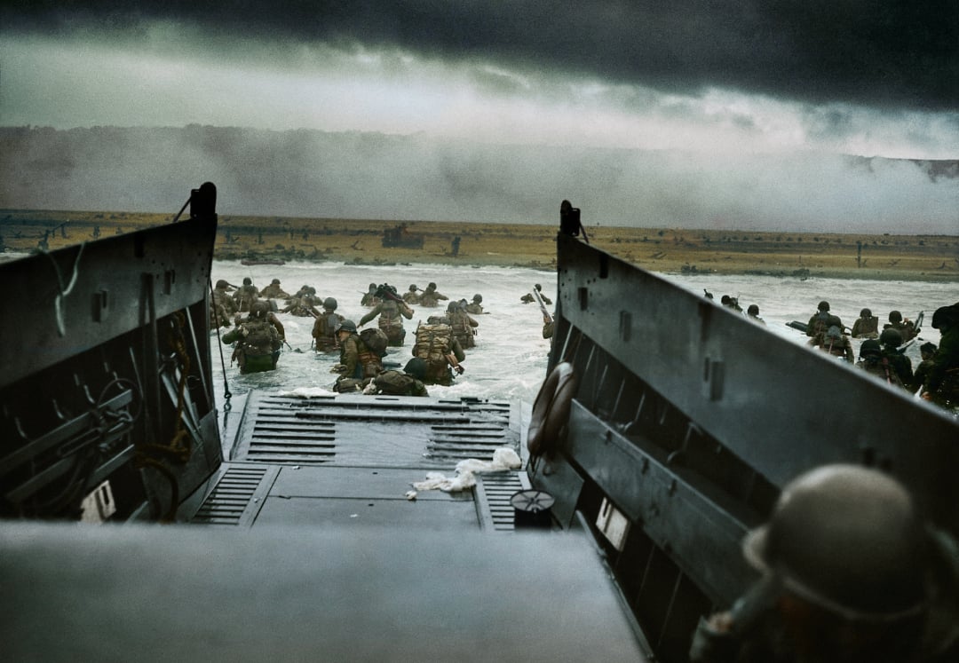 Soldiers wade through water from a landing craft toward a beach under dark, cloudy skies, with smoke and distant cliffs visible in the background. The scene suggests a military invasion or amphibious assault.
