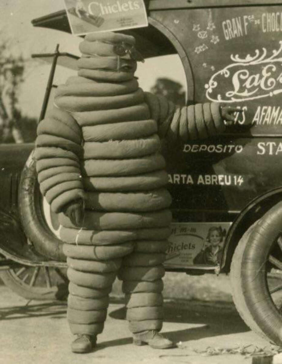A person wearing a vintage Michelin Man costume made of thick, segmented padding stands next to an old car with advertising signs and painted text on its side.