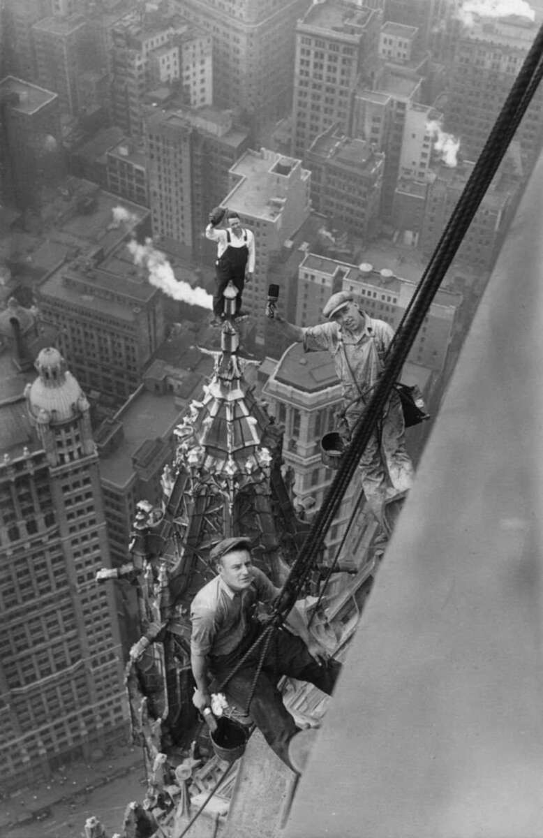 Three construction workers are high up on a skyscraper in a city, working on the building's spire. Tall buildings and city streets are visible far below them, highlighting the extreme height and risk of their job.