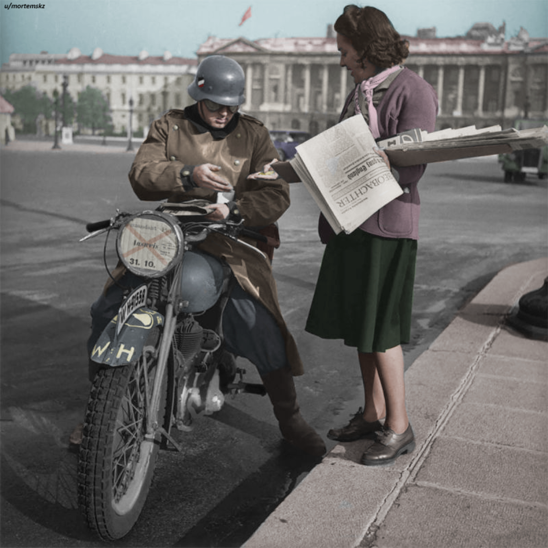 A man in a military coat and helmet sits on a motorcycle, buying a newspaper from a woman holding papers on a city street with large buildings in the background.