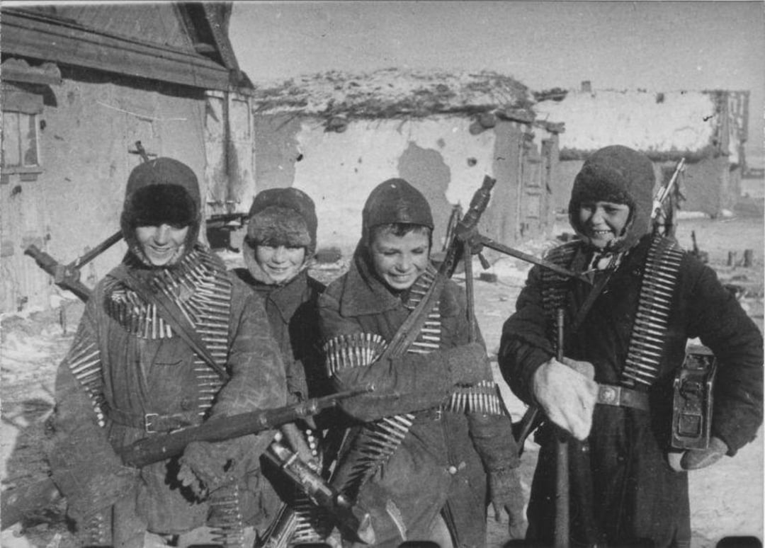 Four young soldiers dressed in winter uniforms and hats stand smiling in the snow, carrying rifles and wearing ammunition belts across their chests. Dilapidated buildings and a snow-covered landscape are in the background.