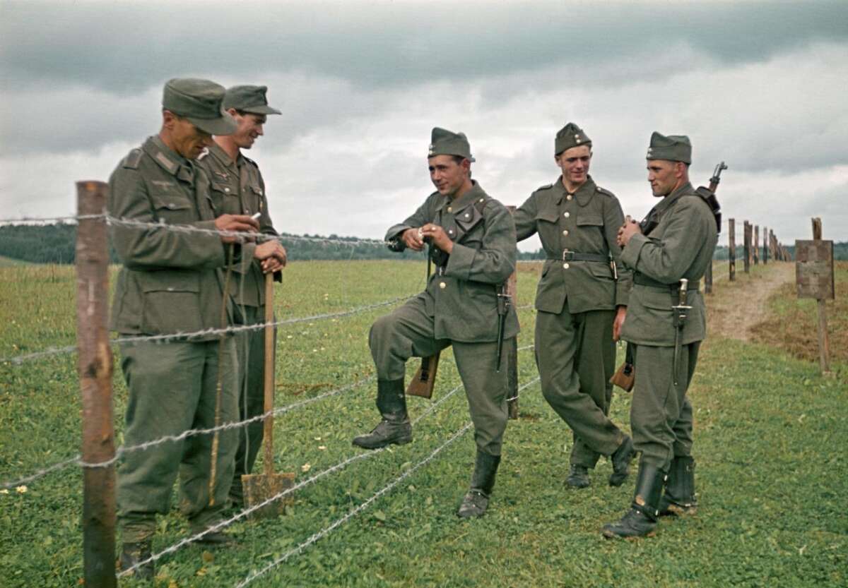 Five soldiers in green uniforms stand by a barbed wire fence in a grassy field, some holding tools and one with a leg propped on the fence, under a cloudy sky.