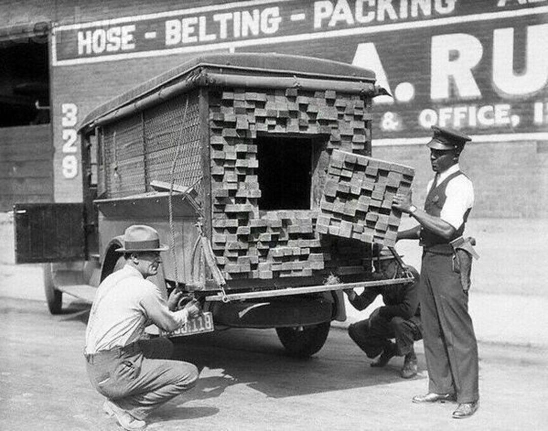 A black-and-white photo shows two men, one in a police uniform, inspecting a truck loaded with bricks; a hidden compartment inside the bricks is revealed by a removed section at the back of the vehicle.