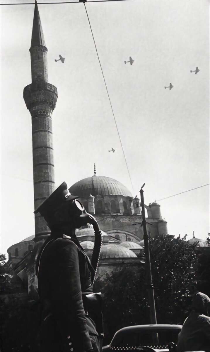 A person in a military uniform and gas mask stands in front of a mosque with a tall minaret, while several airplanes fly overhead against a clear sky. The photo appears to be black and white.