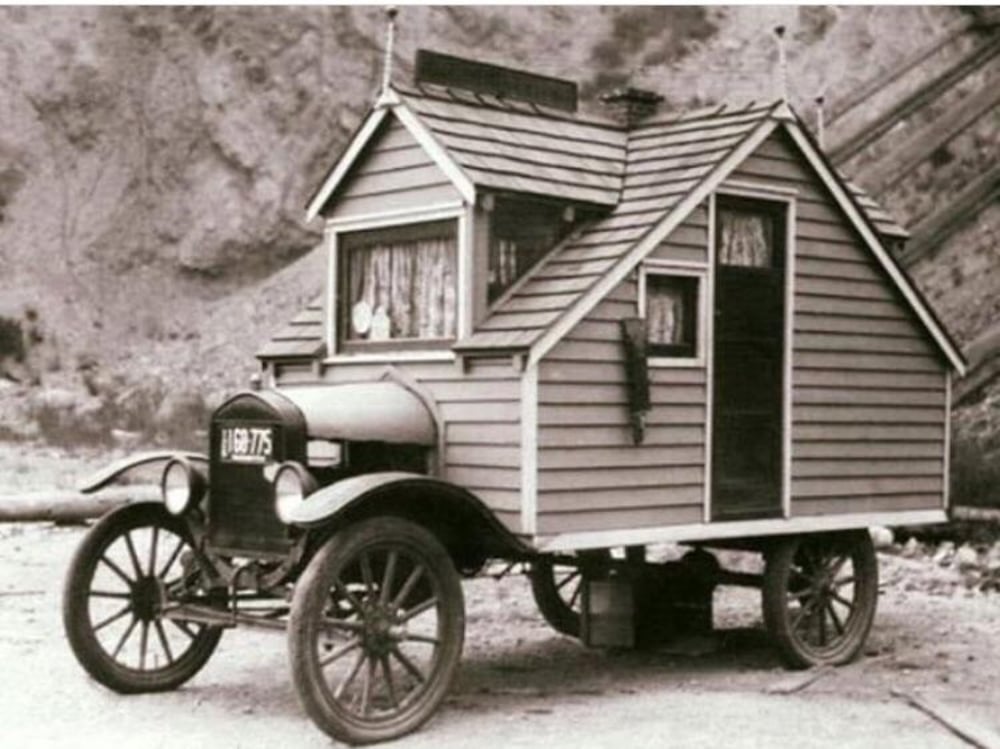 A vintage black-and-white photo shows an old-fashioned car modified with a small wooden house structure built on its chassis, featuring windows with curtains, a door, and a sloped roof.