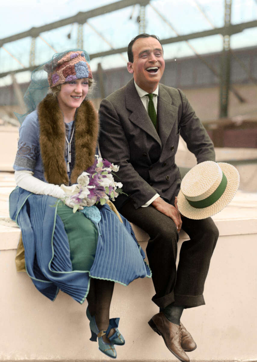 A smiling woman in a blue 1920s dress and hat holding flowers sits next to a laughing man in a suit and tie who holds a straw hat. They sit outdoors on white steps with a metal structure in the background.