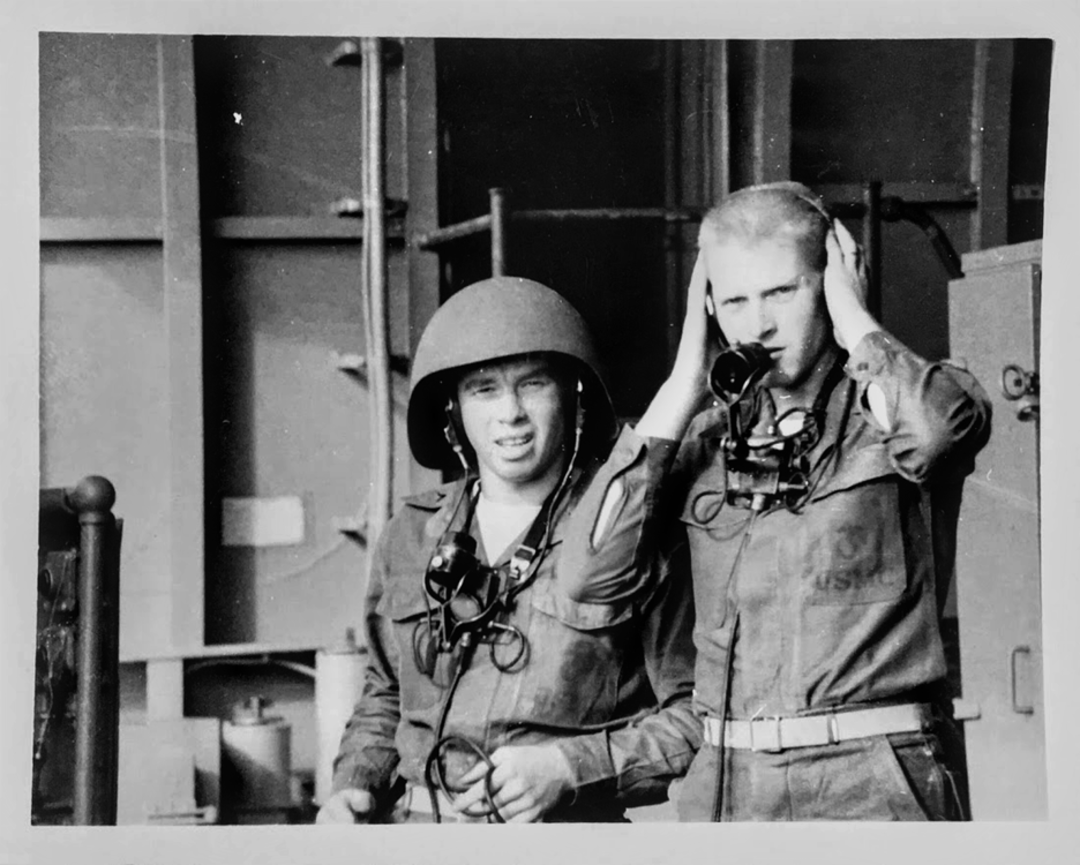 Two men in military uniforms stand side by side; one wears a helmet and the other holds both hands to his ears. Both have headsets and microphones around their necks. Industrial equipment is visible in the background.