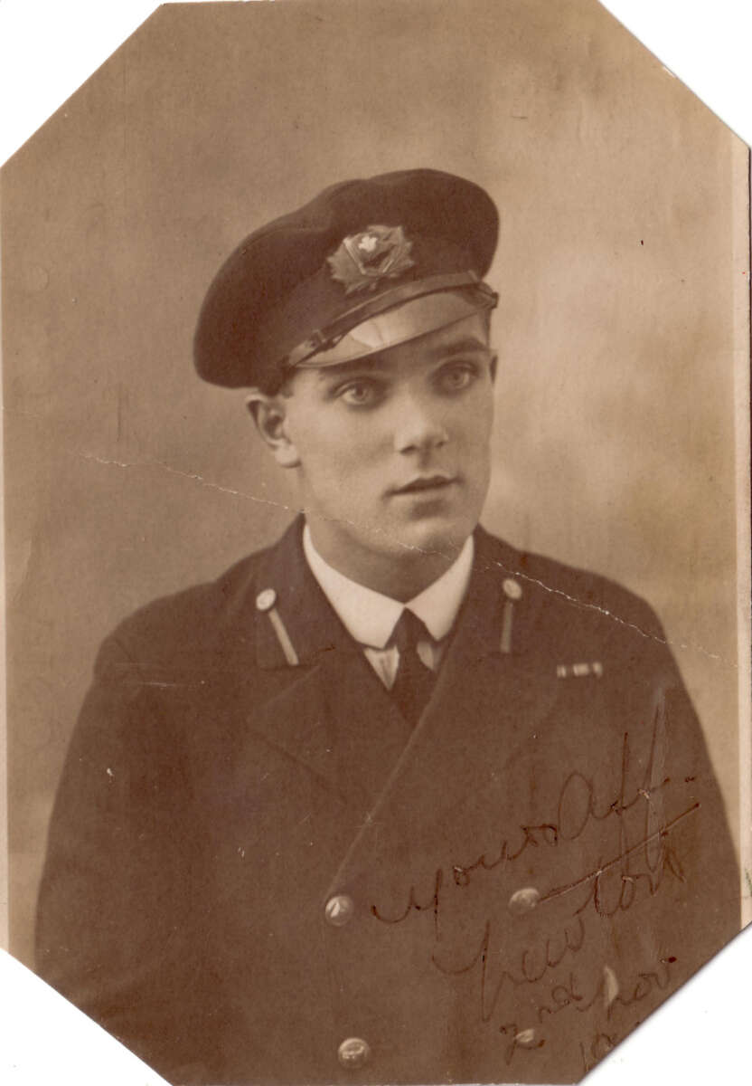 A young man in an old-fashioned military uniform and cap poses for a sepia-toned portrait. His jacket has two rows of buttons and insignia on the collar. The photo has handwriting in the lower right corner.