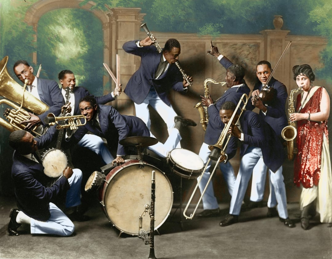 A lively jazz band in blue suits energetically plays brass and percussion instruments, while a woman in a red dress and headpiece stands at the right, holding a saxophone. A painted backdrop of a garden is behind them.