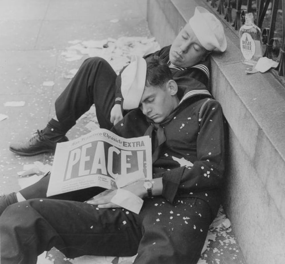 Two sailors in uniform sleep on a city sidewalk amid confetti. One holds a newspaper with the bold headline "PEACE!" while an empty liquor bottle rests nearby, suggesting a celebration.