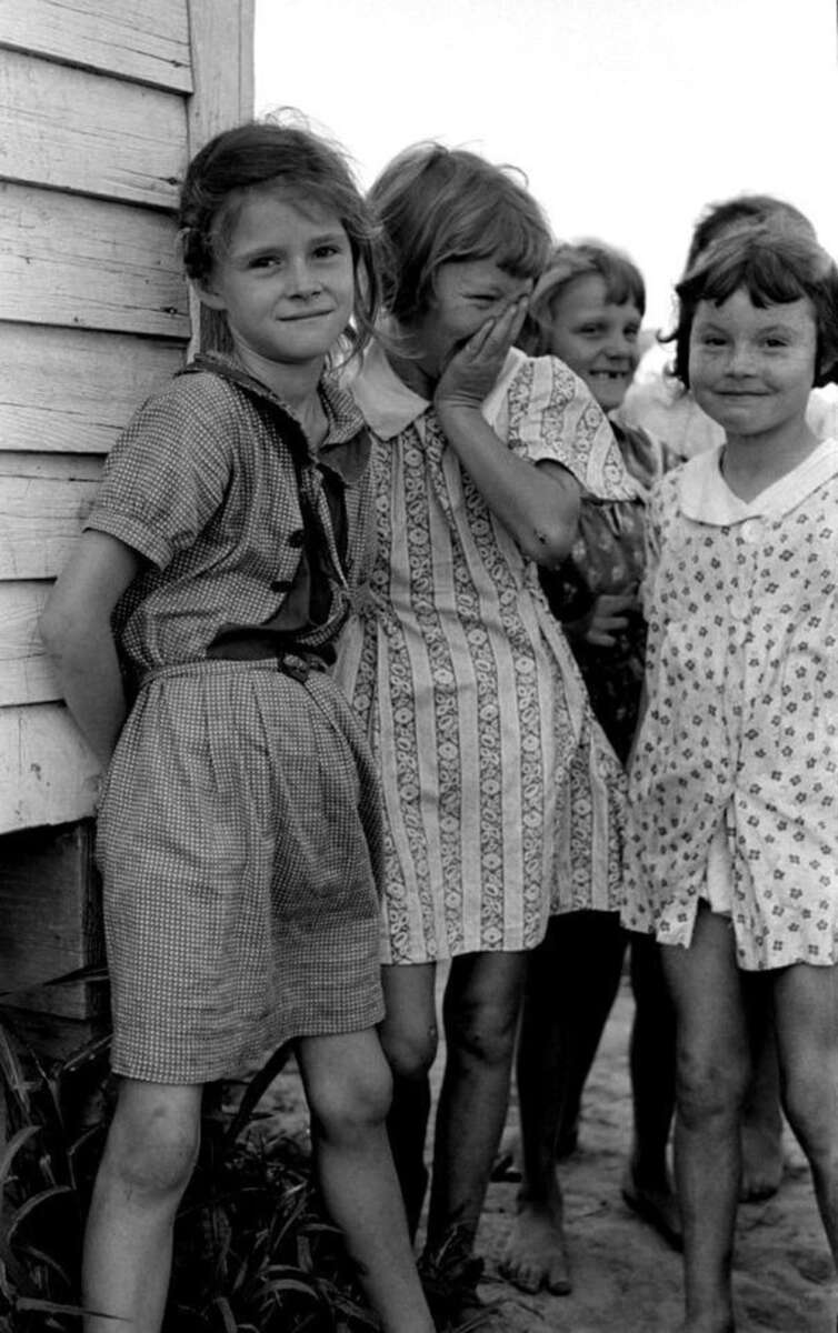 Four young girls stand close together beside a wooden building, smiling and laughing. They wear patterned dresses and appear carefree, with bare feet and tousled hair, suggesting a casual, rural setting.