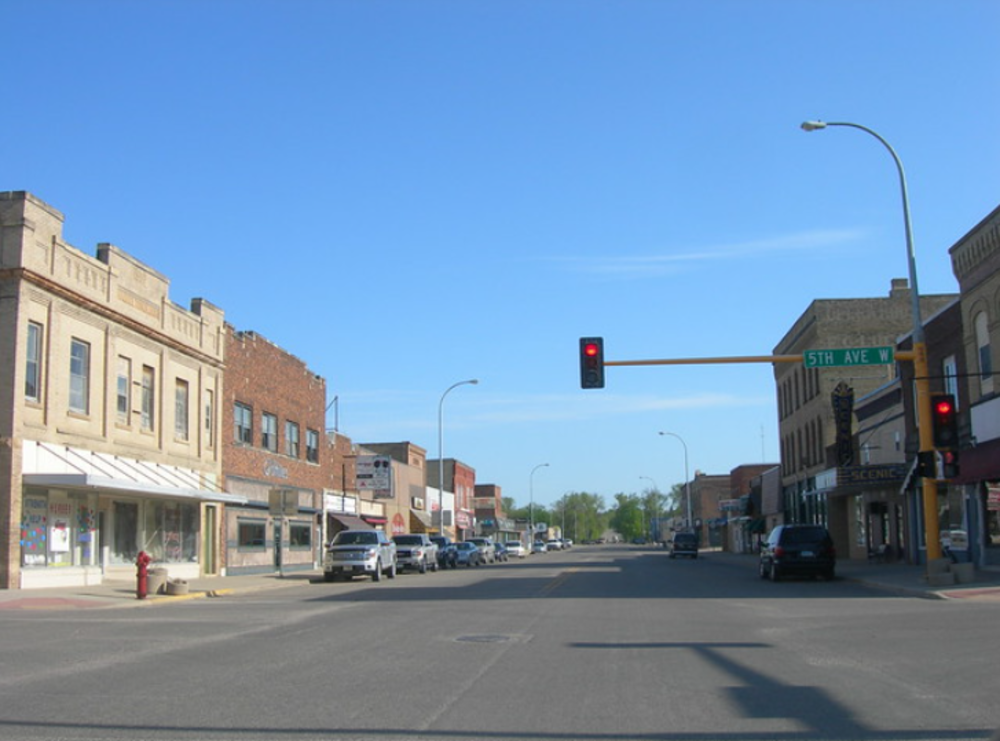 A quiet small-town street with historic brick buildings, parked cars, and a red traffic light at the intersection of 5th Ave under a clear blue sky.