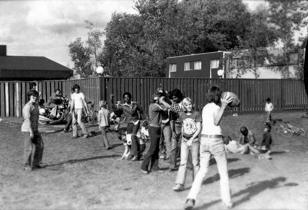 A group of children and teenagers play outside on a grassy area; some stand and talk, while others play with a ball. Bicycles, a motorbike, and buildings are visible in the background on a sunny day.
