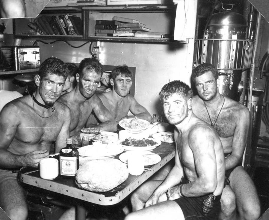 Five shirtless, dirty men sit around a small table covered with food and drinks in a cramped, cluttered room, appearing tired but relaxed, possibly in a submarine or ship setting.