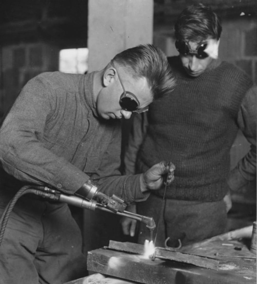 Two men wearing protective goggles work together in a workshop; one is welding metal with a torch while the other observes closely, both focused on the task.