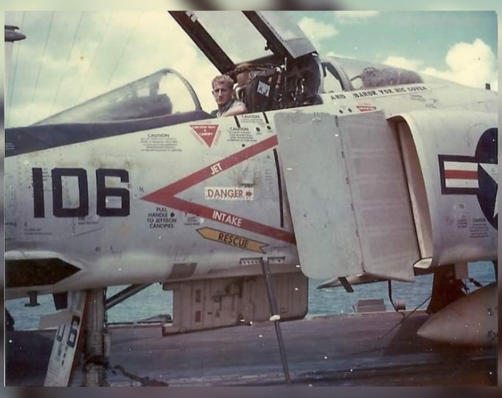 A pilot sits in the open cockpit of a military fighter jet marked with "106" and various warning labels. The aircraft is on the ground, and the sky is partially cloudy in the background.