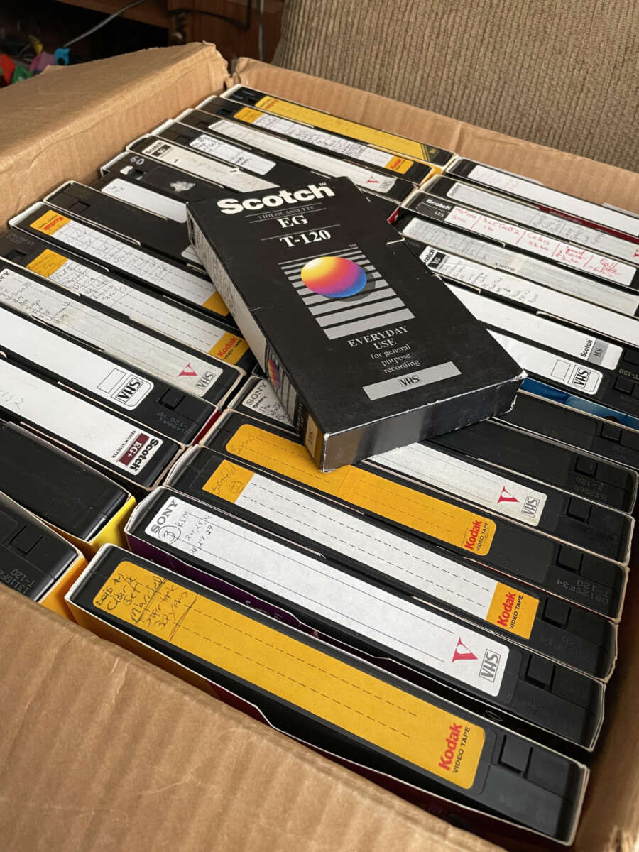 A cardboard box filled with VHS tapes, some labeled with handwritten notes. One Scotch T-120 video cassette is placed on top, and several Kodak-branded tapes are visible among the collection.