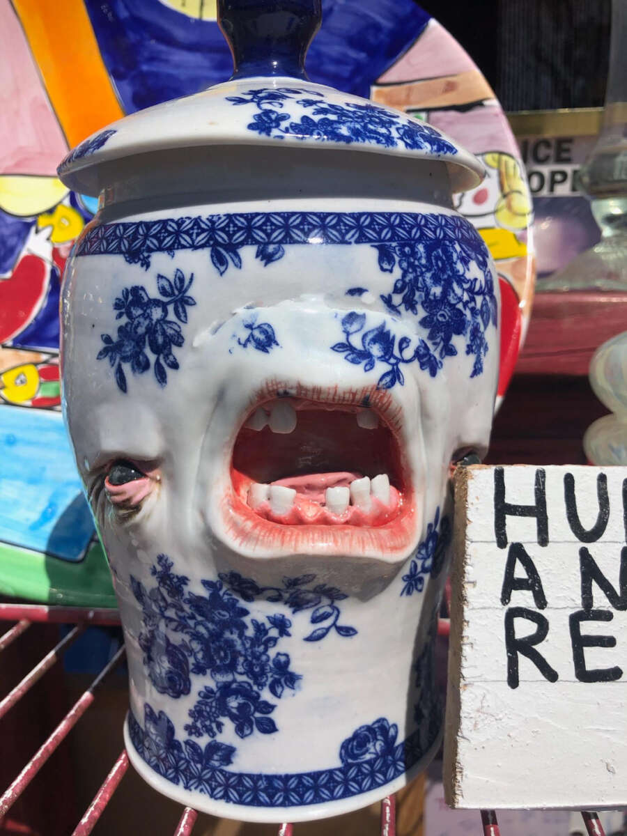 A blue and white porcelain jar with realistic sculpted human facial features, including eyes and an open mouth with teeth, sits on a shelf next to a partially visible sign and colorful plates in the background.