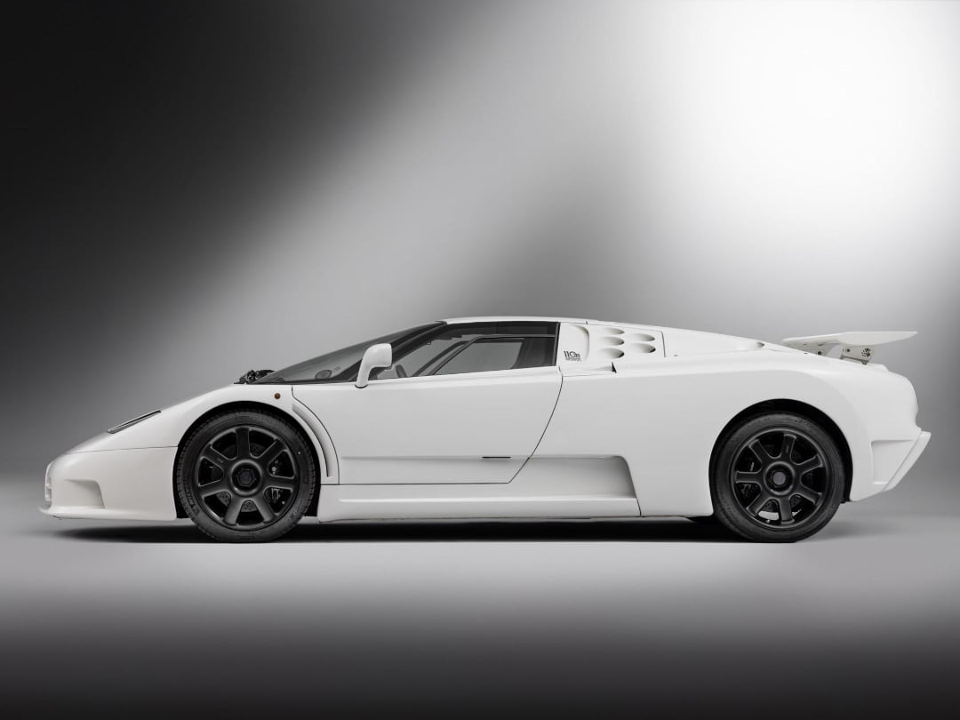 A white sports car with a low, aerodynamic design, black alloy wheels, and a rear spoiler is shown in a studio setting with a gradient gray background.