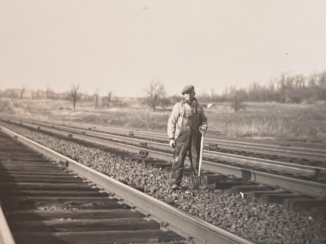 A man wearing overalls and a cap stands on railroad tracks, holding a tool. The scene is outdoors with bare trees and open fields in the background, and the image is in black and white.