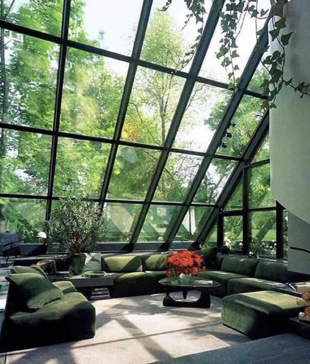 A sunlit living room with floor-to-ceiling angled glass walls, abundant greenery outside, green sofas arranged in a circle, and a coffee table with red flowers in the center.