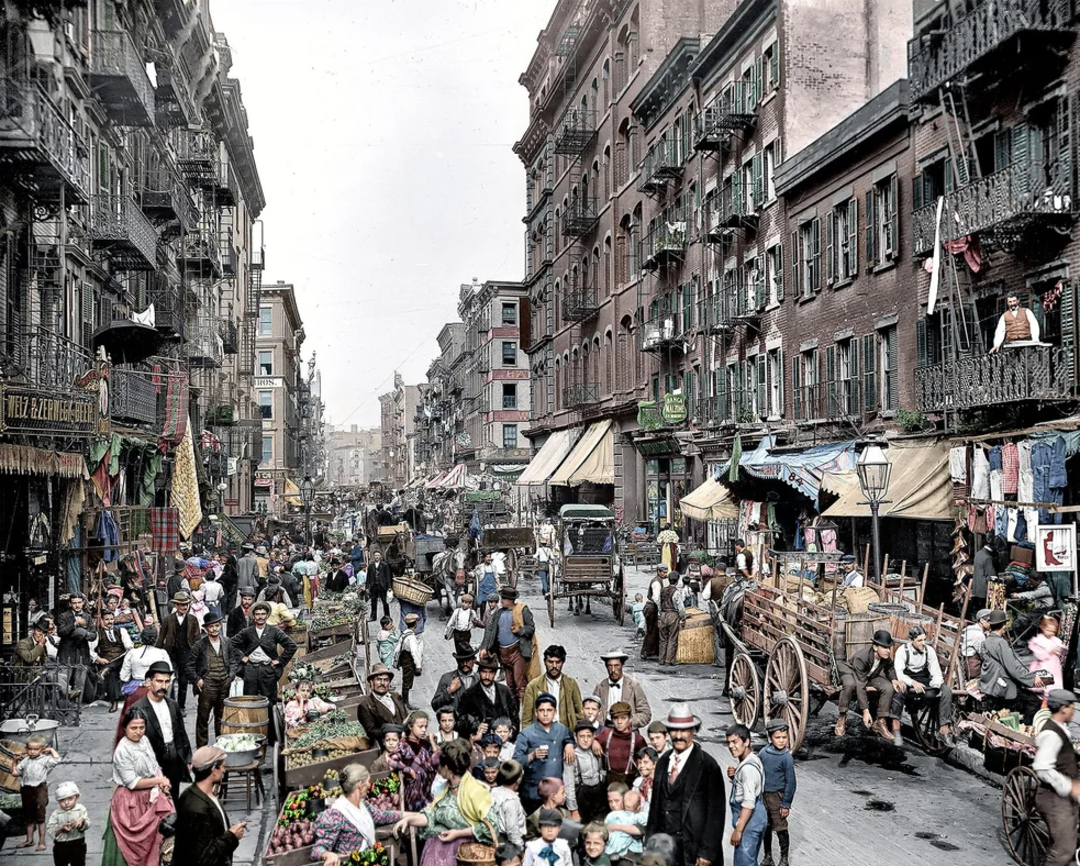 A busy city street scene from the early 1900s shows crowds of people, street vendors with carts, children, horse-drawn wagons, and tall buildings with many balconies and laundry hanging outside.