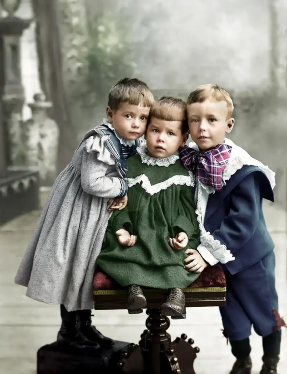 Three young children in vintage clothing pose for a portrait; two stand on either side of a seated child. The boys wear blue outfits and the girl is in a green dress. The image appears colorized and has a soft, historic background.