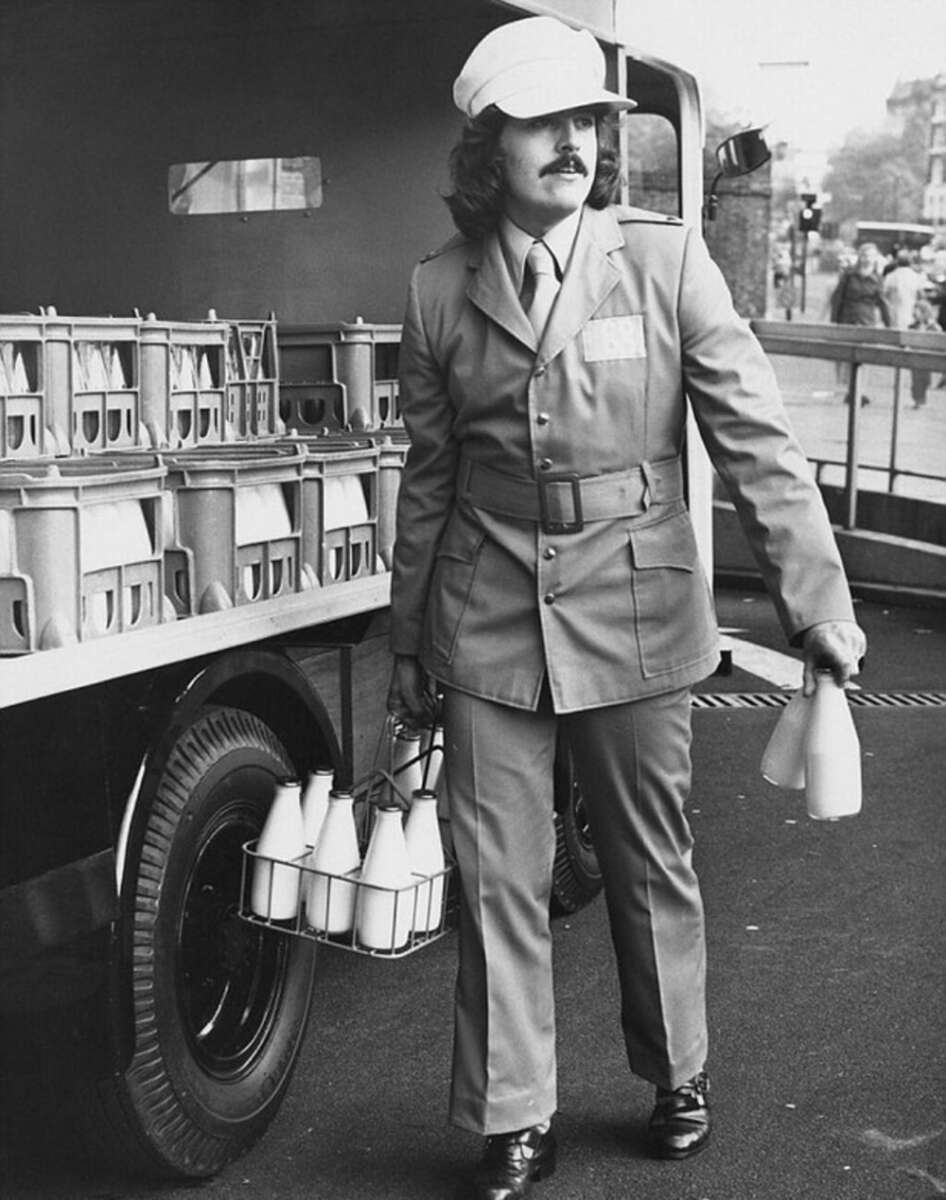 A milk delivery person in a uniform and cap carries glass milk bottles from a truck loaded with crates of milk bottles on a city street. The image is in black and white.