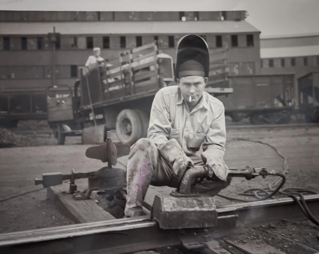 A worker wearing a welding helmet sits on train tracks while welding, with a cigarette in his mouth. Industrial buildings and a truck loaded with wooden planks are visible in the background.