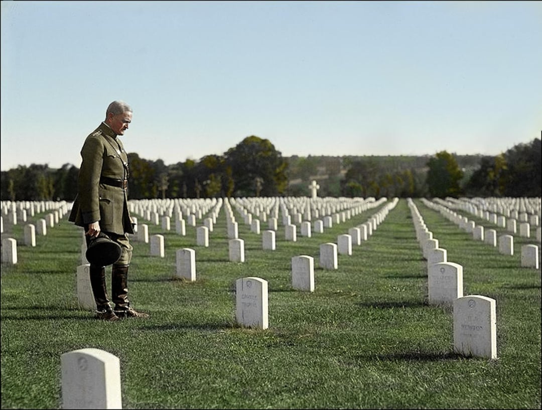 A man in military uniform stands solemnly in a cemetery, holding his hat and looking down at a white gravestone among rows of identical headstones stretching across a grassy field.