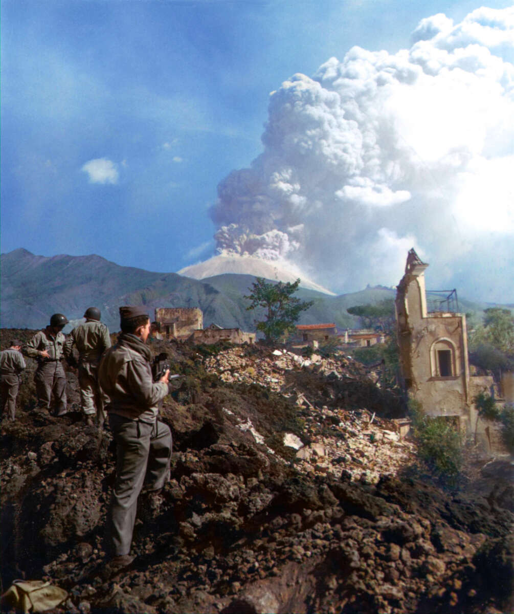 Several soldiers stand on rocky, ash-covered ground, observing a distant volcano erupting with a large plume of smoke. Nearby, ruins of damaged buildings are visible amid the volcanic debris.